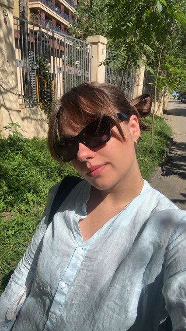 A selfie of a woman with bangs wearing dark sunglasses and a light blue button-up shirt, standing outdoors near a fence and greenery.