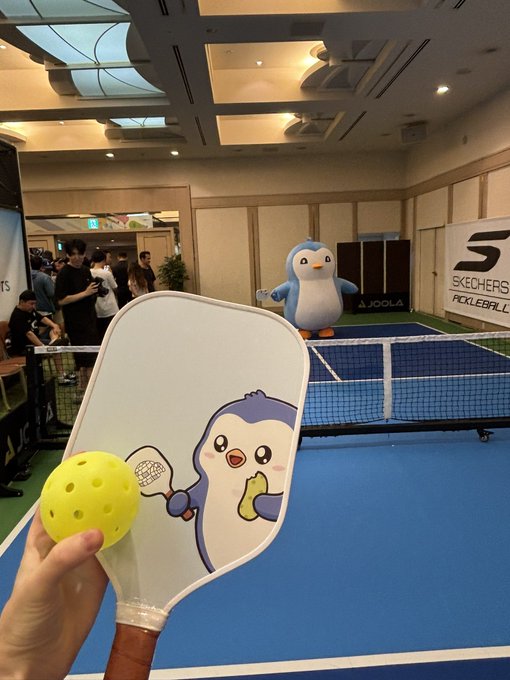 A pickleball player holds up a paddle featuring a cartoon penguin and a yellow wiffle ball, posing across the net from an inflatable penguin mascot on a blue court. The "Skechers Pickleball" and "JOOLA" banners in the background, paired with the face-off framing, playfully set up the post's "Finally a worthy opponent" theme—turning the cute mascot into a tongue-in-cheek rival ready for a match.