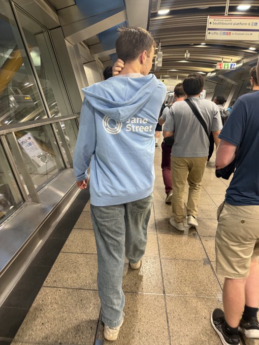 A man in a light blue hoodie with the "Jane Street" logo prominently displayed on the back walks through a subway station, spotted by a passerby. The post humorously critiques quant traders' lack of operational security (opsec) for wearing branded company merch in public, where their employer is instantly identifiable.