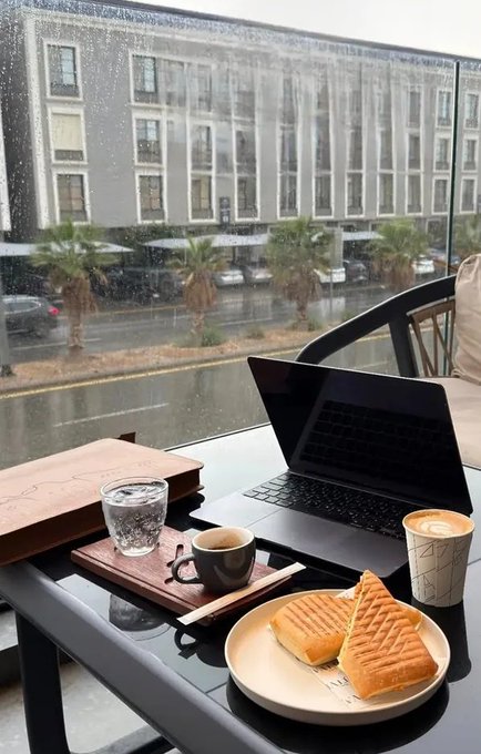 The image shows an open laptop computer sitting on top of a table, with two sandwiches placed next to it. There are also two cups of coffee on the table. A window can be seen behind the laptop, providing a view of a street outside. The scene suggests that someone is working or studying while enjoying their meal and beverages in a comfortable setting.
