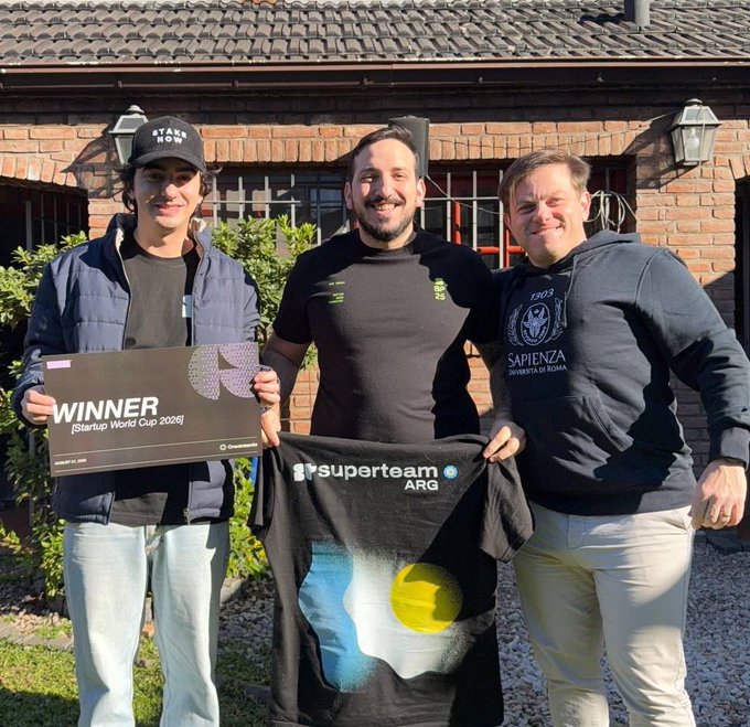 Three men stand smiling in front of a brick house, holding a "WINNER [Startup World Cup 2026]" sign and a "superteam ARG" shirt featuring the Argentine flag design, celebrating their recent victory as noted in the congratulatory post referencing Nora's expansion to Brazil.
