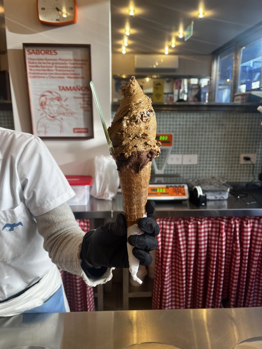 A gloved hand holds up a large, tall waffle cone filled with multiple scoops of gelato, including chocolate and caramel flavors, inside a shop with a menu board on the wall.