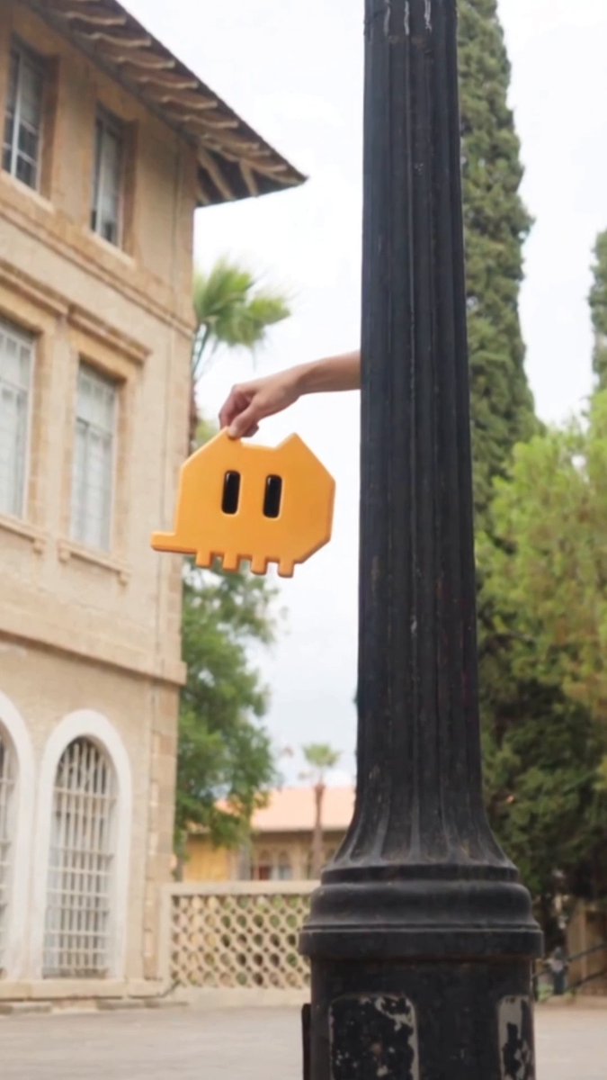 A hand peeks out from behind a lamppost, holding an orange cat-shaped sign to surprise the viewer—illustrating the "always appearing at the right time" theme of the Bybit AI ad, which invites users to chat daily for a chance to win an iPhone via i.bybit.com/1kabgJy7.