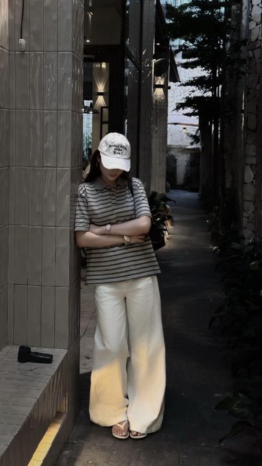 A person wearing a white baseball cap, striped t-shirt, and white wide-leg pants stands with arms crossed against a tiled wall in a dimly lit outdoor alleyway, with a small dumbbell visible on a ledge beside them.