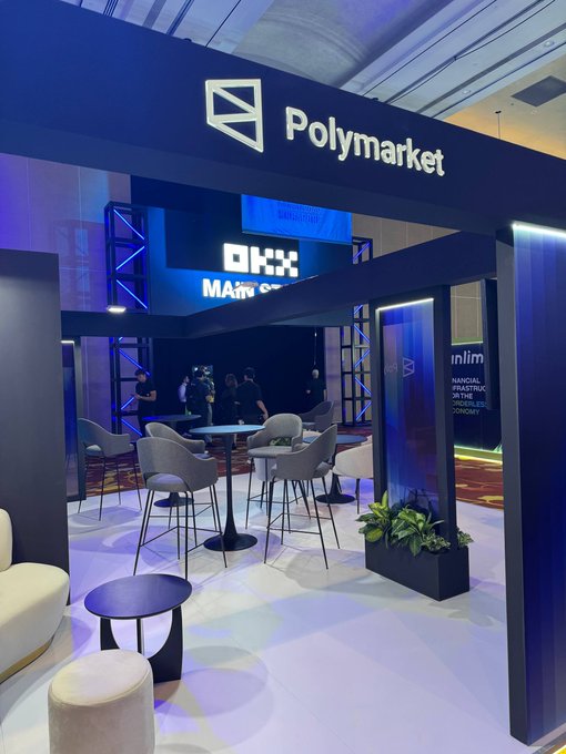 A lounge area at an event featuring a 'Polymarket' sign above a seating space with chairs, tables, and an OKX sign visible in the background.