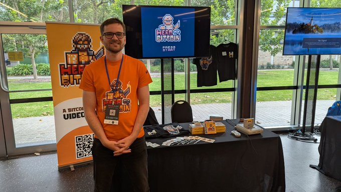 A man in an orange t-shirt stands at a 'Head2Head Stream' promotional booth displaying merchandise, t-shirts, and a screen with the event logo.
