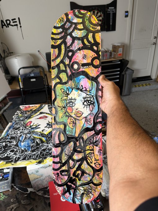 A hand holds up a skateboard deck covered in vibrant, chaotic artwork featuring a central character with large eyes surrounded by abstract patterns and faces.