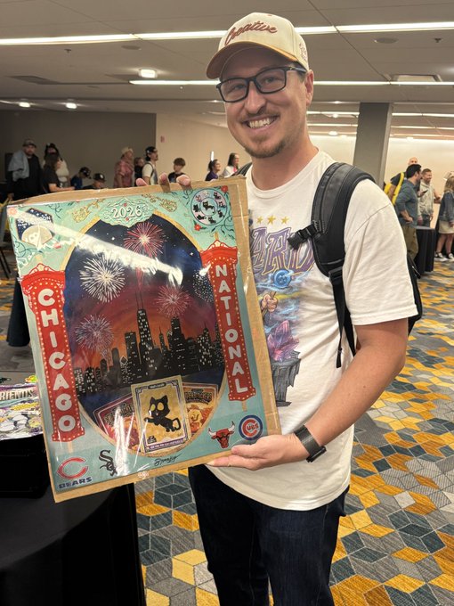 A smiling man wearing a cap and glasses holds a 2025 Chicago Cubs poster featuring a Chicago skyline, fireworks, and various team imagery in a convention hall setting.