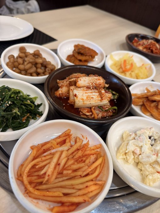 A spread of Korean side dishes (banchan) including seasoned greens, kimchi, soybeans, sweet potato fries, and what appears to be spicy fish or tofu stew, fuel the preparation for the Solana Summit Korea in a nod to the hearty meals shared at crypto industry events.