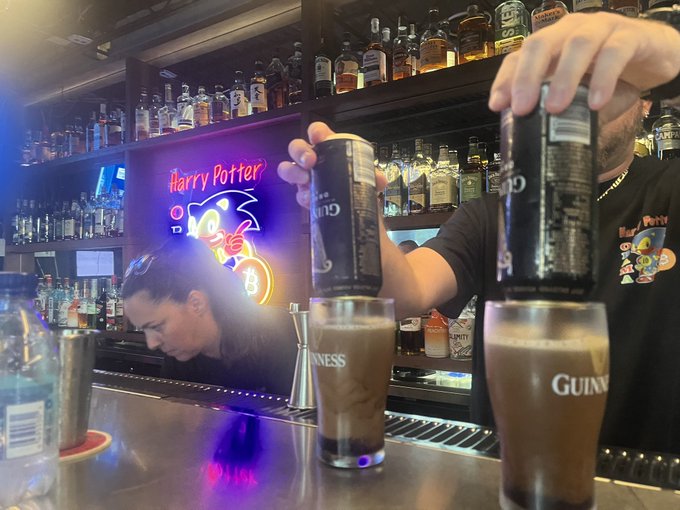 Two bartenders at a 'Harry Potter' themed bar pour Guinness beers from cans into branded glasses.