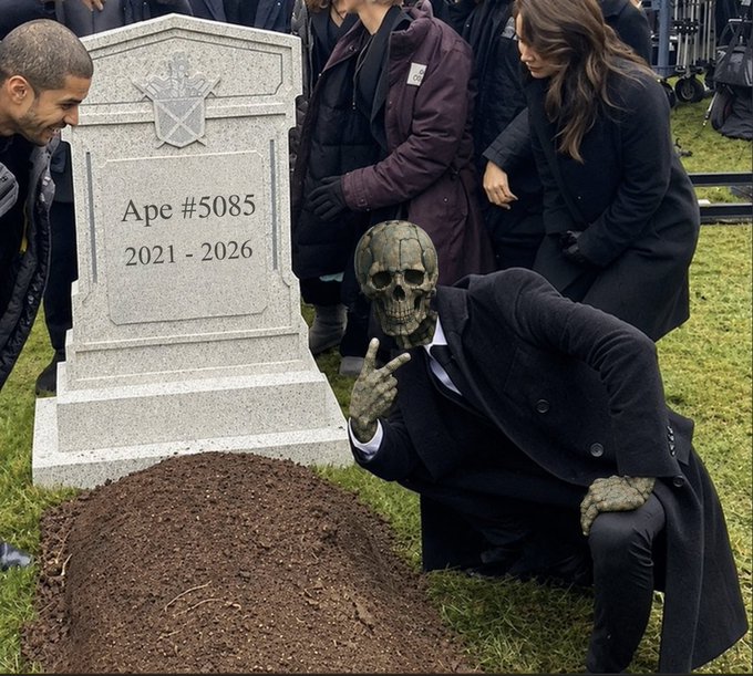 A surreal scene depicts a figure with a skull head wearing a black suit giving a peace sign in front of a tombstone marked "Ape #5085, 2021 - 2026," with mourners gathered around.