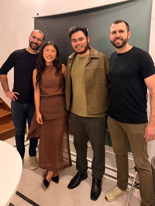 Four people pose together for a photo indoors against a green chalkboard; they are dressed in casual-to-smart attire, including a woman in a brown dress and men in dark and olive-toned clothing.