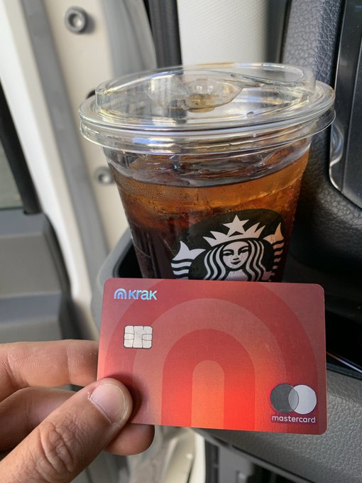 A hand holds a Kraken Mastercard debit card in front of an iced Starbucks coffee inside what appears to be an airplane or train.