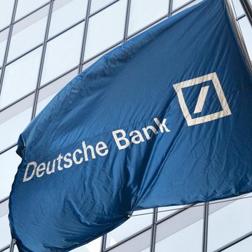 A blue flag bearing the white text 'Deutsche Bank' and its slash-in-square logo waving in front of a modern glass building.