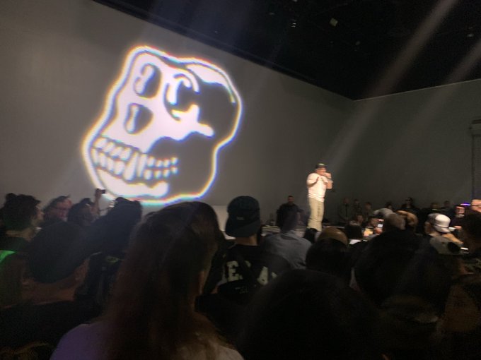 A speaker addressing a crowd at an event with a large, colorful projection of a skull on the screen behind them.
