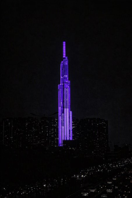 A nighttime cityscape featuring a tall skyscraper illuminated in purple against a dark sky, with smaller lit buildings visible below.