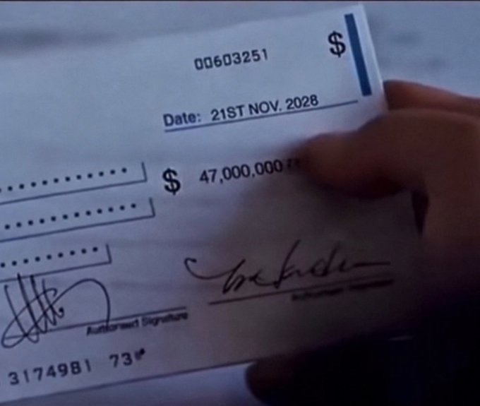 A close-up of a hand holding a bank check dated "21ST NOV. 2028" made out for "$ 47,000,000" with check number "00603251" and visible signatures.