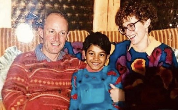 A vintage family photograph showing a smiling man in a red patterned sweater, a young boy in a blue sweater, and a smiling woman with curly hair and glasses in a patterned sweater, posed together on a striped couch.