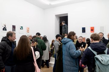 Crowded gallery viewers gather around the framed generative artwork "hello world" by qubibien, displayed at Galerie Met in Berlin during Art on Tezos, with small framed pieces visible on the white walls as attendees socialize and engage with the exhibition.