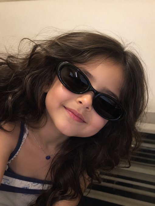 Close-up selfie of a young woman with long brown hair wearing black oval sunglasses and a delicate necklace, with a soft smile.