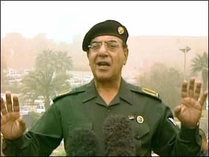 A low-resolution gif or video frame shows a military officer in a green uniform and beret with glasses, speaking with both hands raised, standing in front of microphones with a desert background.