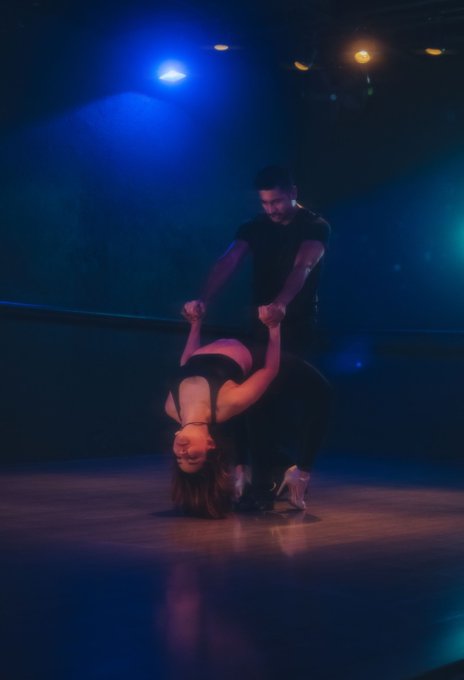 A man and woman perform a dramatic dance lift on a dimly lit stage, with the man holding the woman as she bends backward in a flexible pose, illuminated by moody blue and warm spotlights—symbolizing trust, balance, and seizing the moment, much like the post's message about seizing opportunities before time runs out.