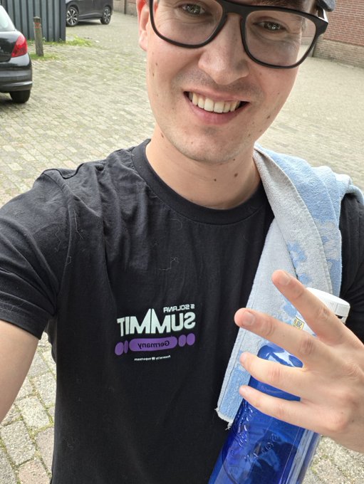 A smiling young man wearing glasses takes a selfie while holding a blue water bottle and a striped towel over his shoulder; he wears a black 'SUMMIT' t-shirt (logo appears mirrored).