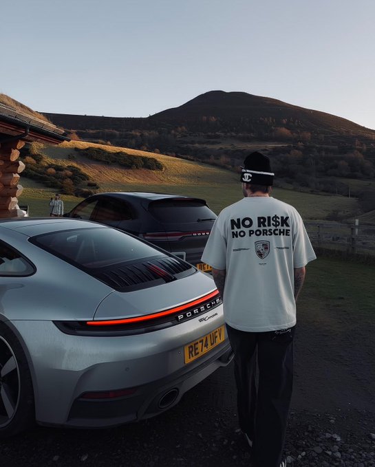 A person wearing a white t-shirt reading 'NO RISK NO PORSCHE' stands with their back to the camera, looking toward a silver Porsche 911 Carrera with a yellow license plate 'RE74 UFP' against a scenic mountain backdrop.