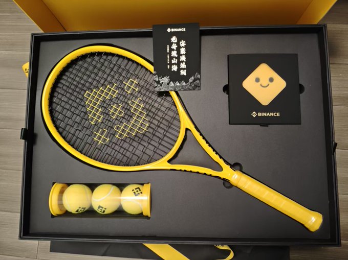 A yellow tennis racket branded with the Binance logo (a diamond shape with a smiley face) displayed in a black gift box with three yellow tennis balls and Binance-branded cards featuring Chinese text.