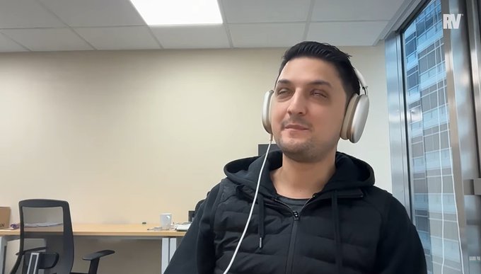 A man wearing a black puffer jacket and headphones sits in an office setting with an 'RV' watermark visible in the corner.