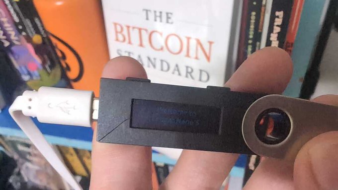 A faded Ledger Nano S hardware wallet is held up in front of a copy of "The Bitcoin Standard," illustrating the post's claim that the device's screen naturally fades with age rather than indicating any tampering or security breach.