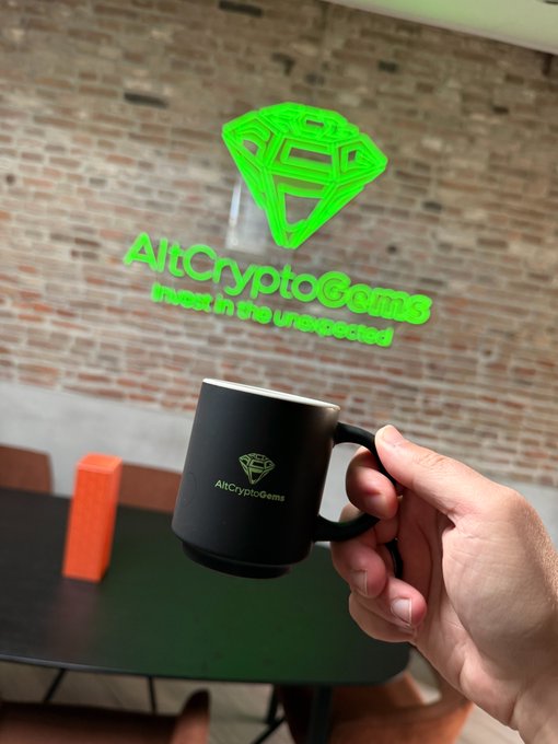 A hand holds a black mug with 'AltCryptoGems' and a diamond logo, with the same logo displayed as a green neon-style overlay and the tagline 'invest in the unexpected' against a brick wall background.