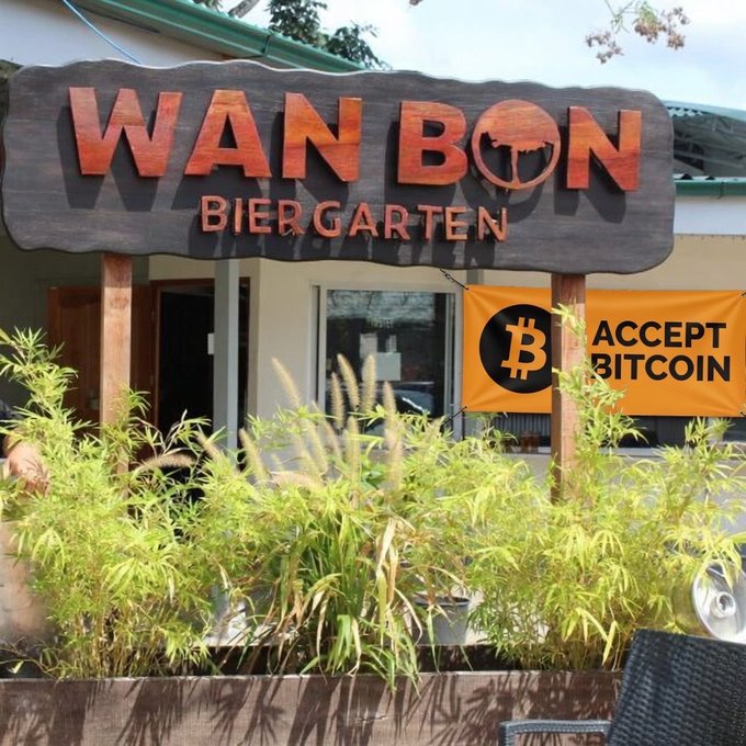 Exterior of Wan Bon Biergarten restaurant featuring a wooden sign and a bright orange banner stating 'ACCEPT BITCOIN' with the Bitcoin logo.