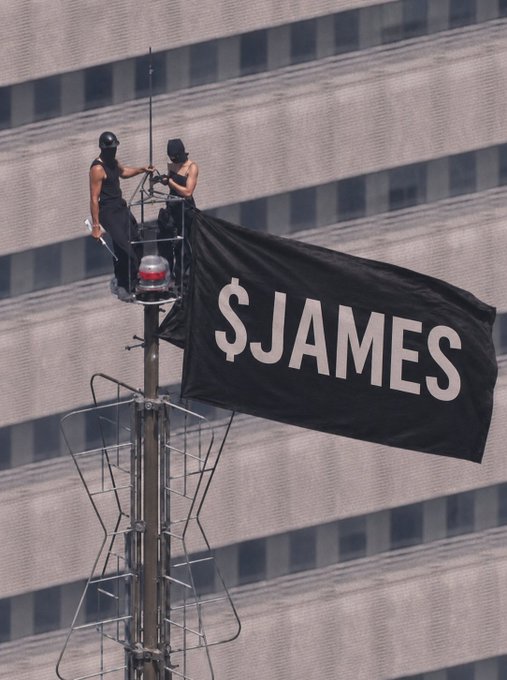 Two people in black balaclavas and tank tops standing on a platform attached to a pole, next to a large black flag reading '$JAMES' in white letters, with a building in the background.