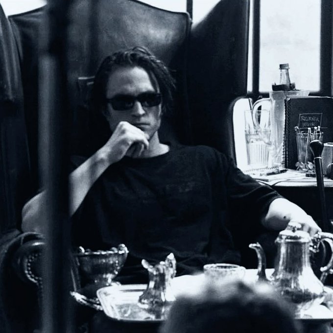 The image features a man sitting in a chair, wearing sunglasses and a black shirt. He has his hand over his mouth as if he's deep in thought or contemplation. The scene takes place around him, with various objects such as cups, bowls, and bottles scattered on the table. There is also another person partially visible at the bottom right corner of the image.

The post that accompanies this image reads: "$neet at ?". This could be a social media post or a message from someone who wants to share their thoughts about the man sitting in the chair, possibly related to his appearance or behavior. The focus on the man's sunglasses and the overall atmosphere of the scene suggests that it might be an attempt to make fun of him or provoke a reaction from others.