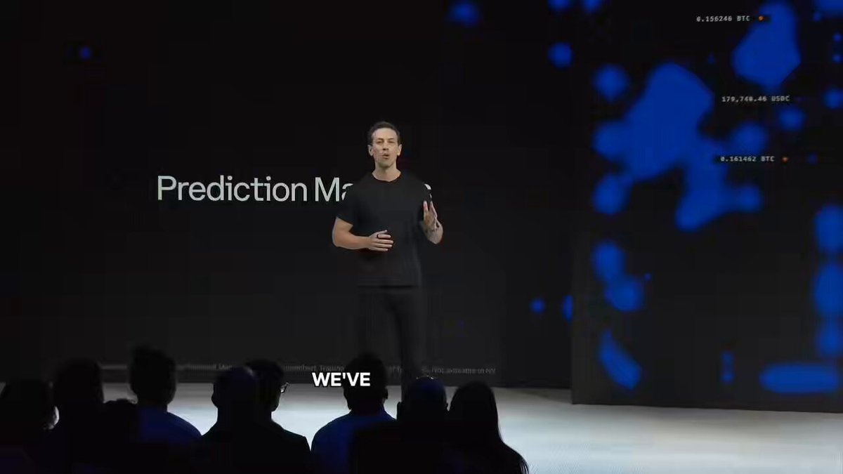 The image shows a man standing at a podium with a microphone, giving a speech or presentation. The background of the image features a large screen displaying blue dots, which could be part of an interactive display or a projection screen for visual aids during the event. The caption below the image reads "Prediction Mr.: We've got it!".