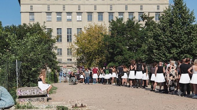 A line of people, many wearing matching white skirts and black tops, standing outside a large building in what appears to be a queue.