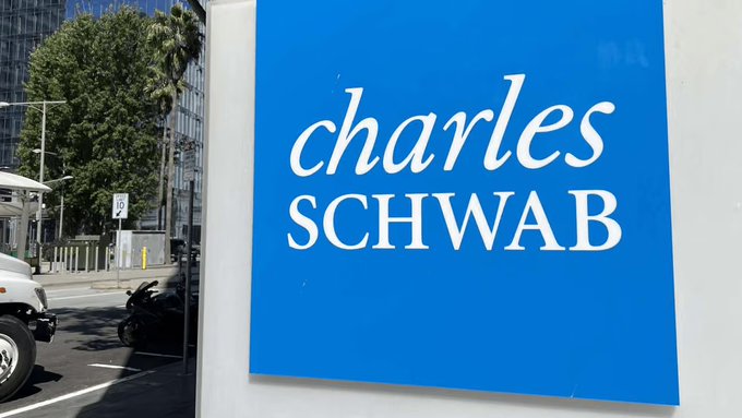 A blue sign displaying the "Charles Schwab" logo is mounted on a white exterior wall, with a street scene featuring a parked motorcycle and trees visible in the background. The image relates to the news that Charles Schwab has launched Bitcoin and Ethereum trading services for its 40 million retail clients, highlighting the financial firm's move into cryptocurrency markets.