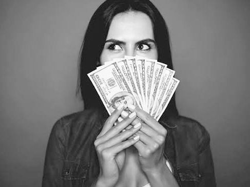 A black and white photo of a woman holding a fan of US dollar bills in front of her face while looking sideways.