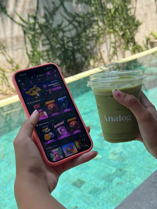 A person's hands hold a smartphone displaying a gaming app interface in one hand and a clear plastic cup of green iced matcha drink labeled "Analog" in the other, with a pool in the background.