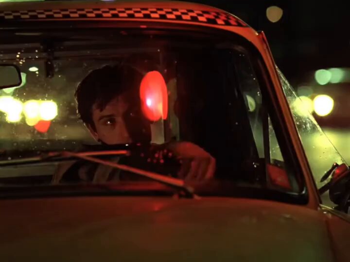 The image shows a man sitting inside a taxi cab at night, looking out of the window as he drives down the street. The car's interior light is turned on, illuminating the scene outside. There are also red lights visible in the background, possibly indicating that the driver is using them to signal or communicate with other drivers or pedestrians.