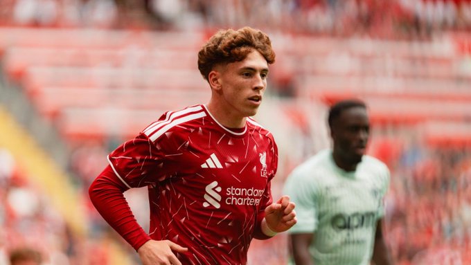 A young Liverpool FC player with curly hair is captured mid-action wearing a red jersey featuring the Adidas logo and "Standard Chartered" sponsor text, while an opponent in a green kit is visible in the background during what appears to be a pre-season friendly match.
