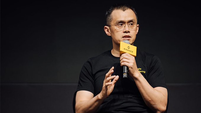 A man wearing glasses and a black t-shirt speaks into a yellow 'BINANCE' microphone while on stage.