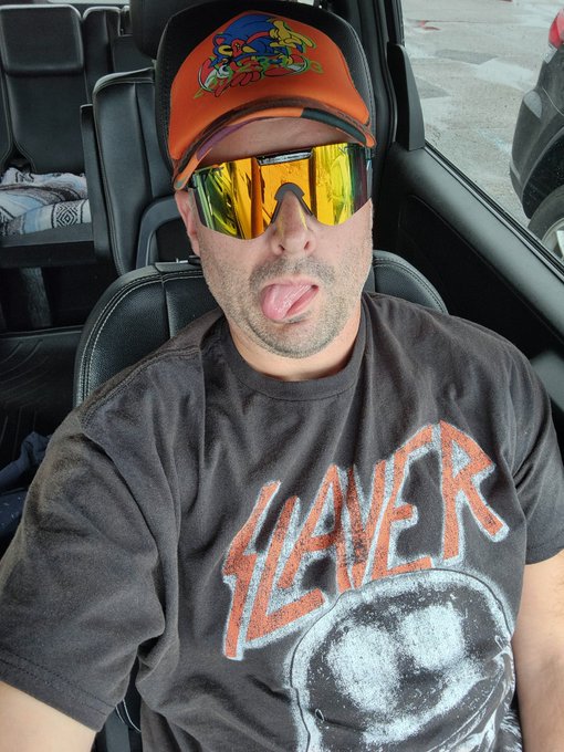 A man in a vehicle wearing a Slayer t-shirt, an orange Tropical Storm cap, and reflective gold sunglasses makes a silly face at the camera.