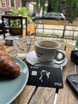The image shows a wooden table with various items, including a cup of coffee, a pastry, and a Visa card. The coffee cup has a white saucer underneath it, while the pastry is placed next to the card. A cell phone can also be seen on the table. The scene suggests that someone might have just finished their breakfast or is about to enjoy one. The presence of the Visa card indicates that they may have recently made a purchase using this card and are now taking a moment to relax with their coffee and pastry before starting their day.