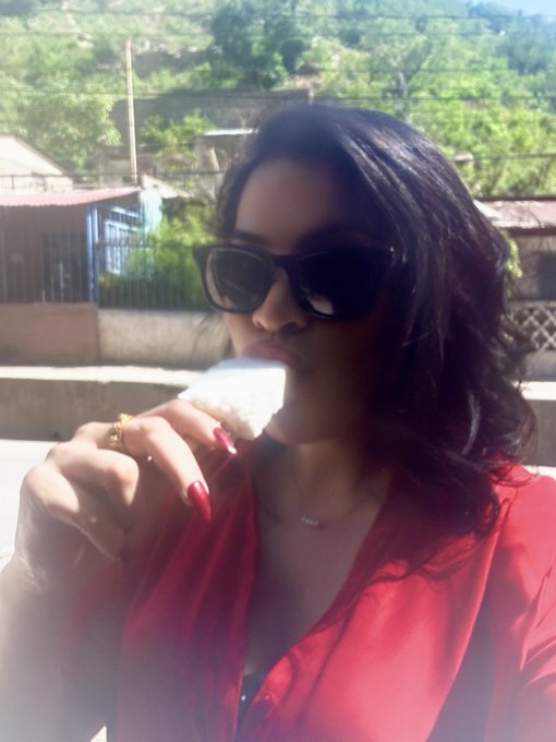 A woman wearing sunglasses and a red shirt holds a piece of food near her mouth in an outdoor setting.