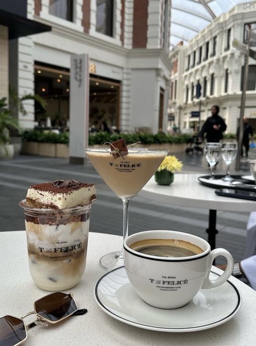 The image shows a table with three cups of coffee and two desserts placed on it. The desserts include a cupcake, a mousse, and a cake. There are also sunglasses resting on the table. The scene takes place outdoors, possibly at an outdoor cafe or restaurant.