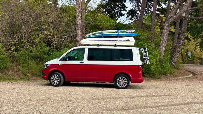 A two-tone red and white Volkswagen camper van parked on a gravel area with surfboards mounted on the roof rack, set against a backdrop of green trees.