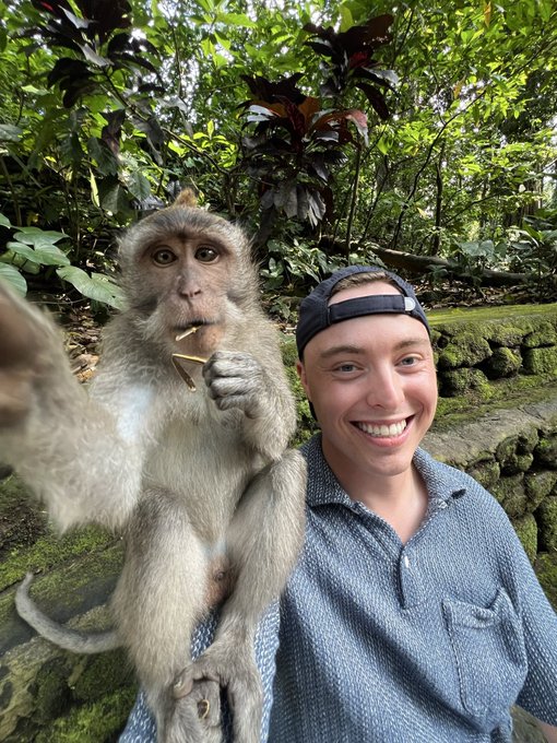A smiling young man wearing a blue button-up shirt and backwards cap poses for a selfie with a macaque monkey holding what appears to be a piece of food in a lush jungle setting.