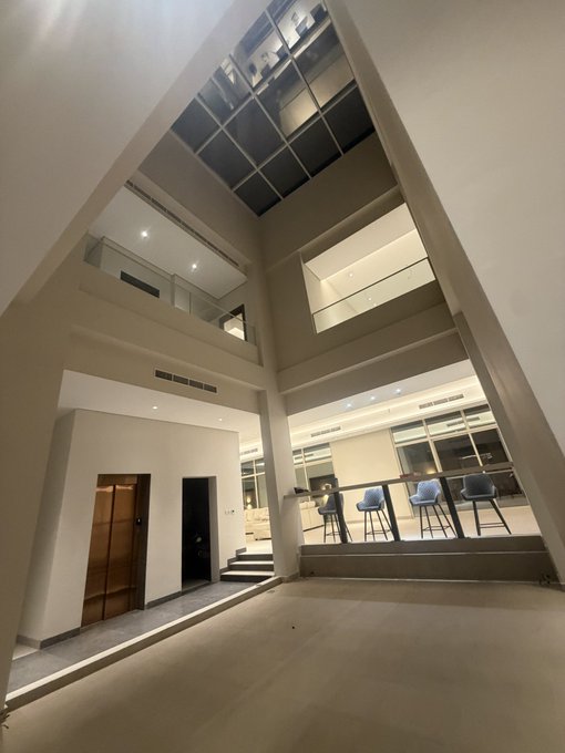 A spacious, double-height room with soaring ceilings, sleek white walls, and a raised bar area with three blue stools showcases dramatic architectural angles still mid-renovation. The reference to hanging a car from the ceiling plays on the cavernous ceiling height, which provides plenty of room for such an ambitious finishing touch.